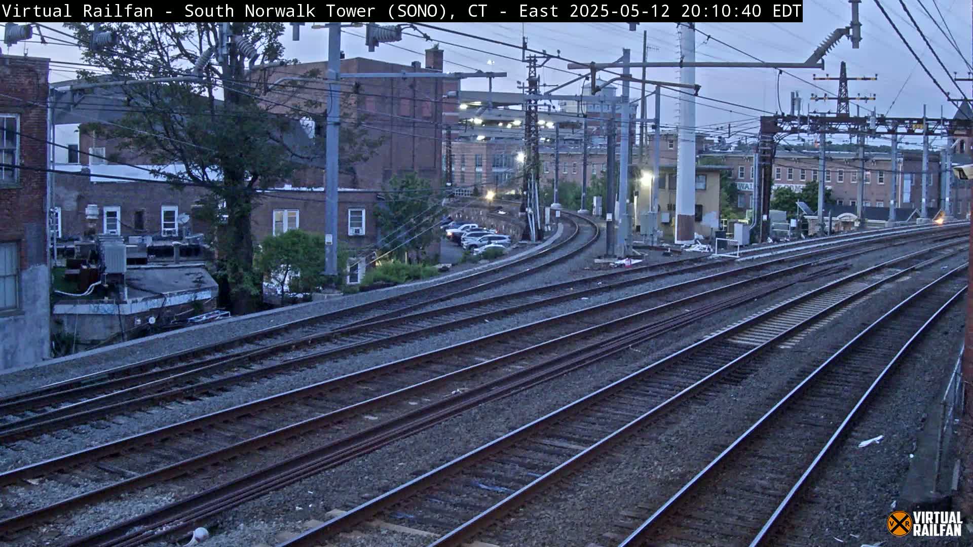 Several sets of train tracks curve gently past buildings under a twilight sky.