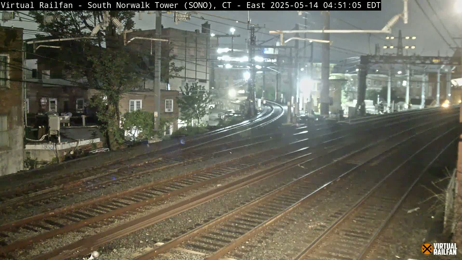 A nighttime view of several train tracks curving away from the camera, passing alongside residential buildings under a dark, possibly slightly rainy sky.