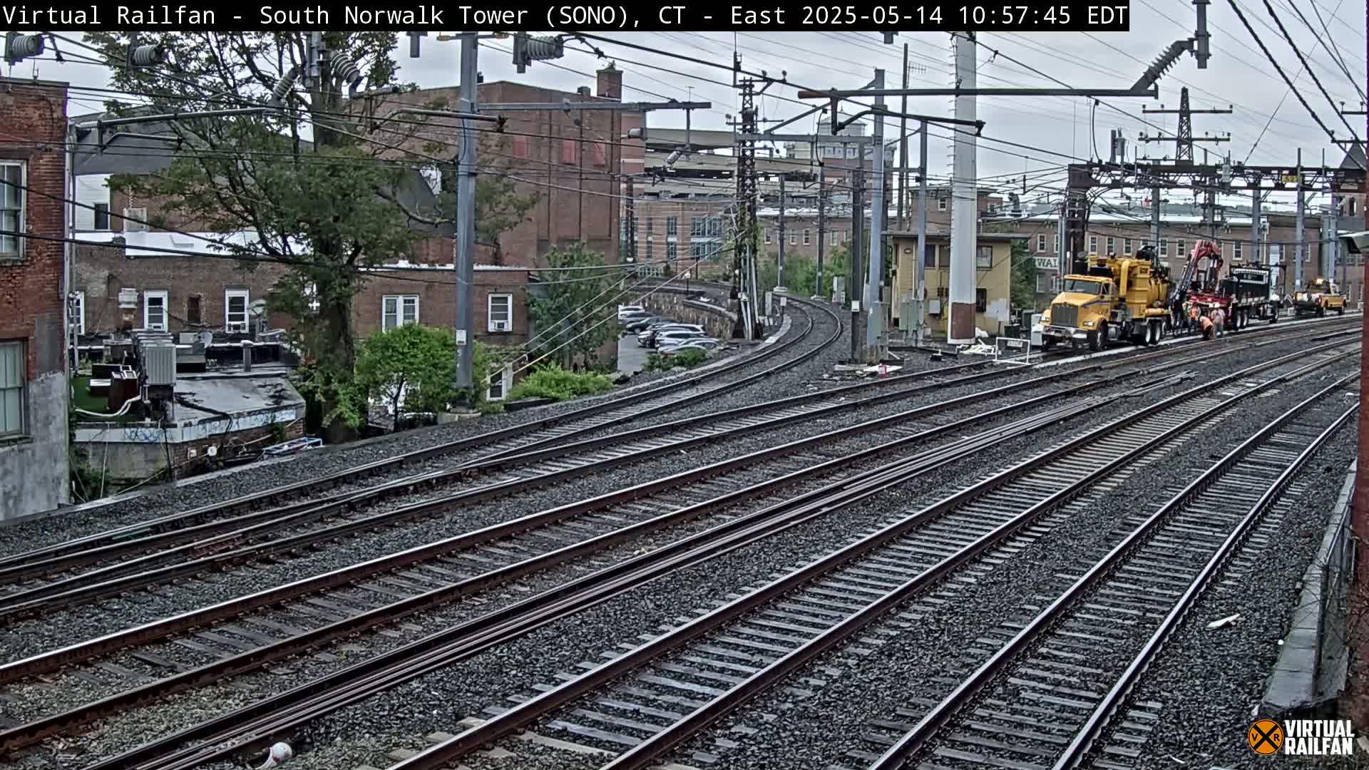A view of several sets of train tracks curving away from the camera, past a yellow piece of railway maintenance equipment and several buildings on an overcast day.