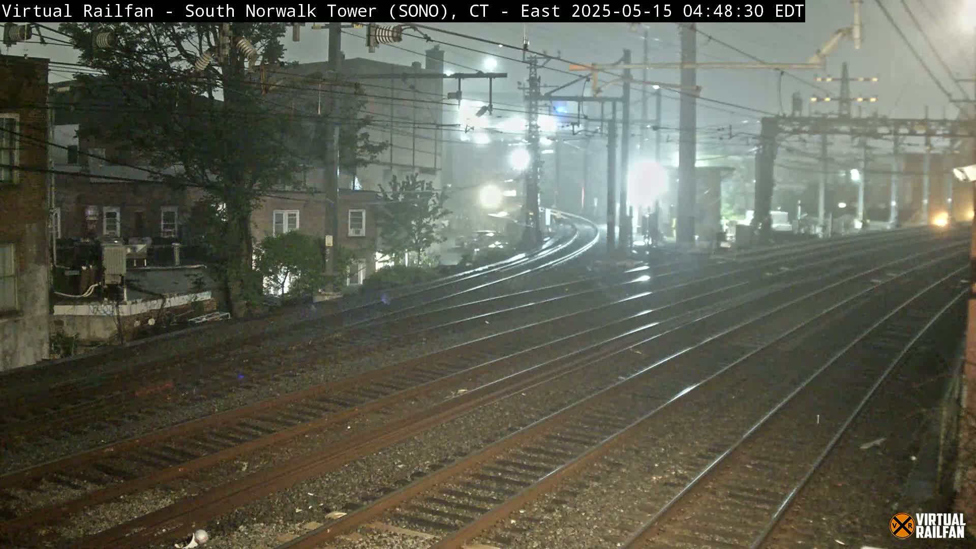 Multiple train tracks curve into the distance under a foggy, nighttime sky, passing alongside buildings and vegetation.