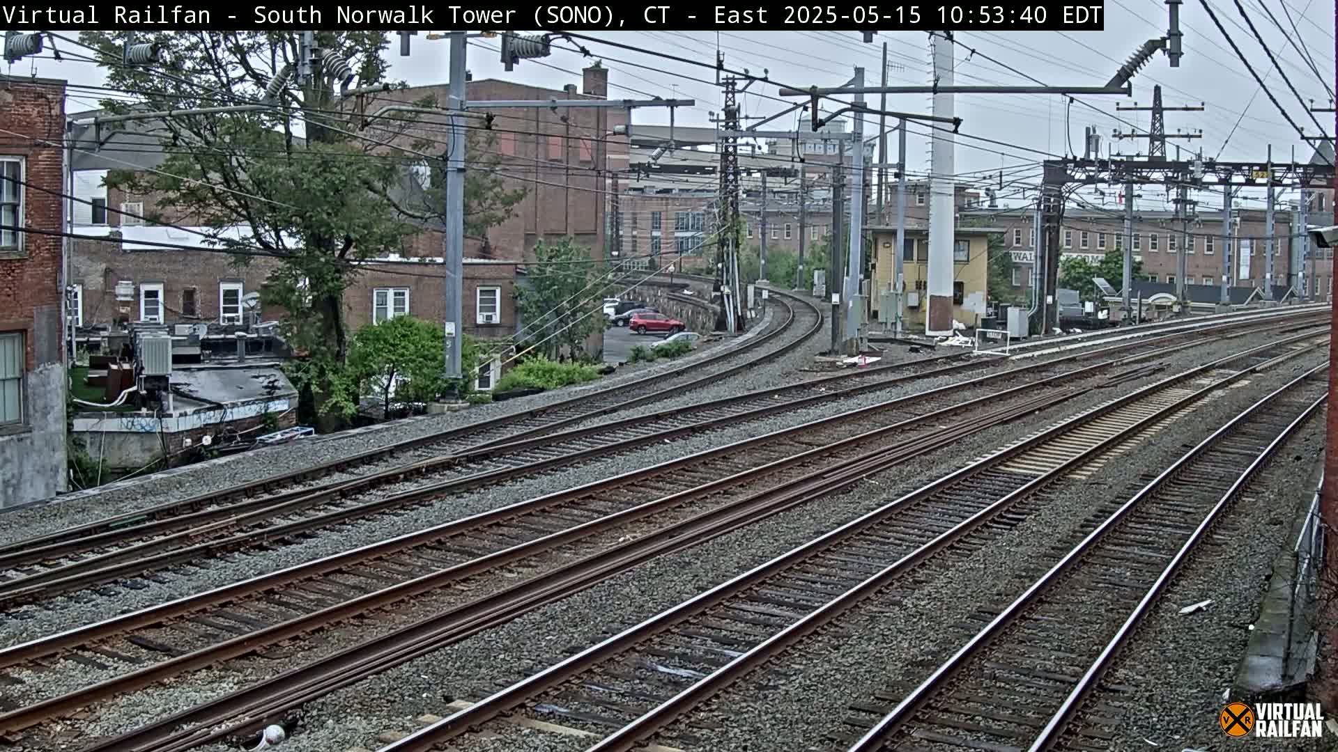 Multiple train tracks curve through an overcast urban area, passing near brick buildings and some greenery.