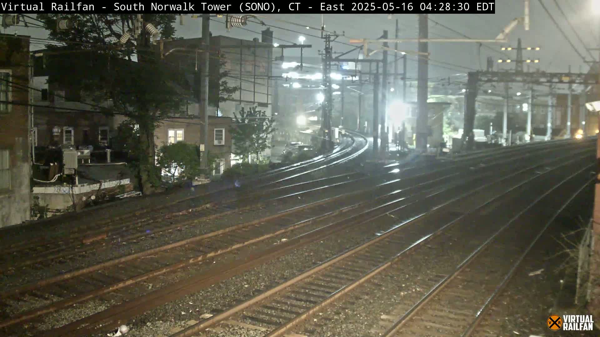 Wet railroad tracks curve away from a nighttime city scene under dark, overcast skies.