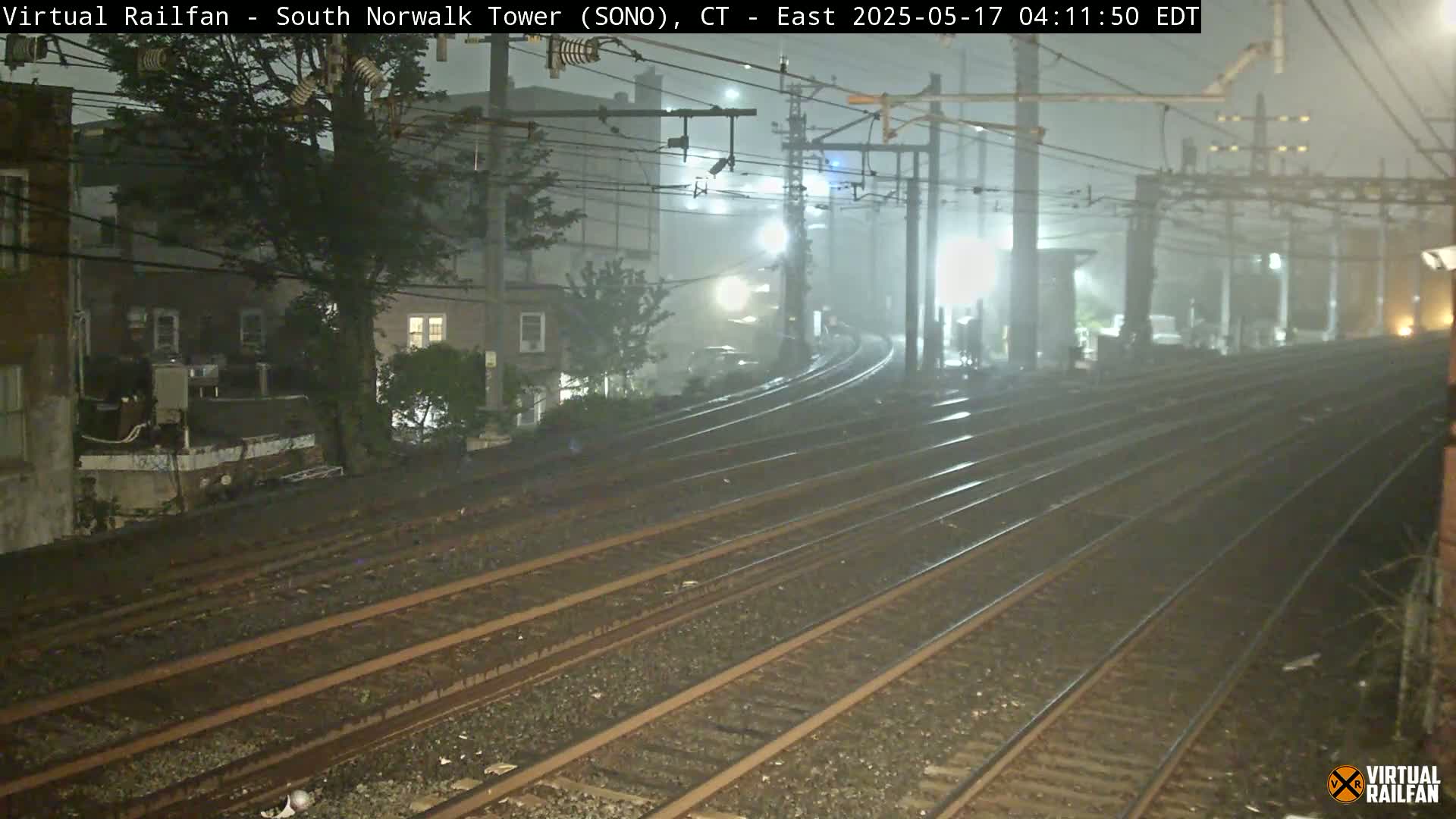 A nighttime view of a foggy train yard with multiple sets of tracks curving away from the camera, bordered by buildings and trees.