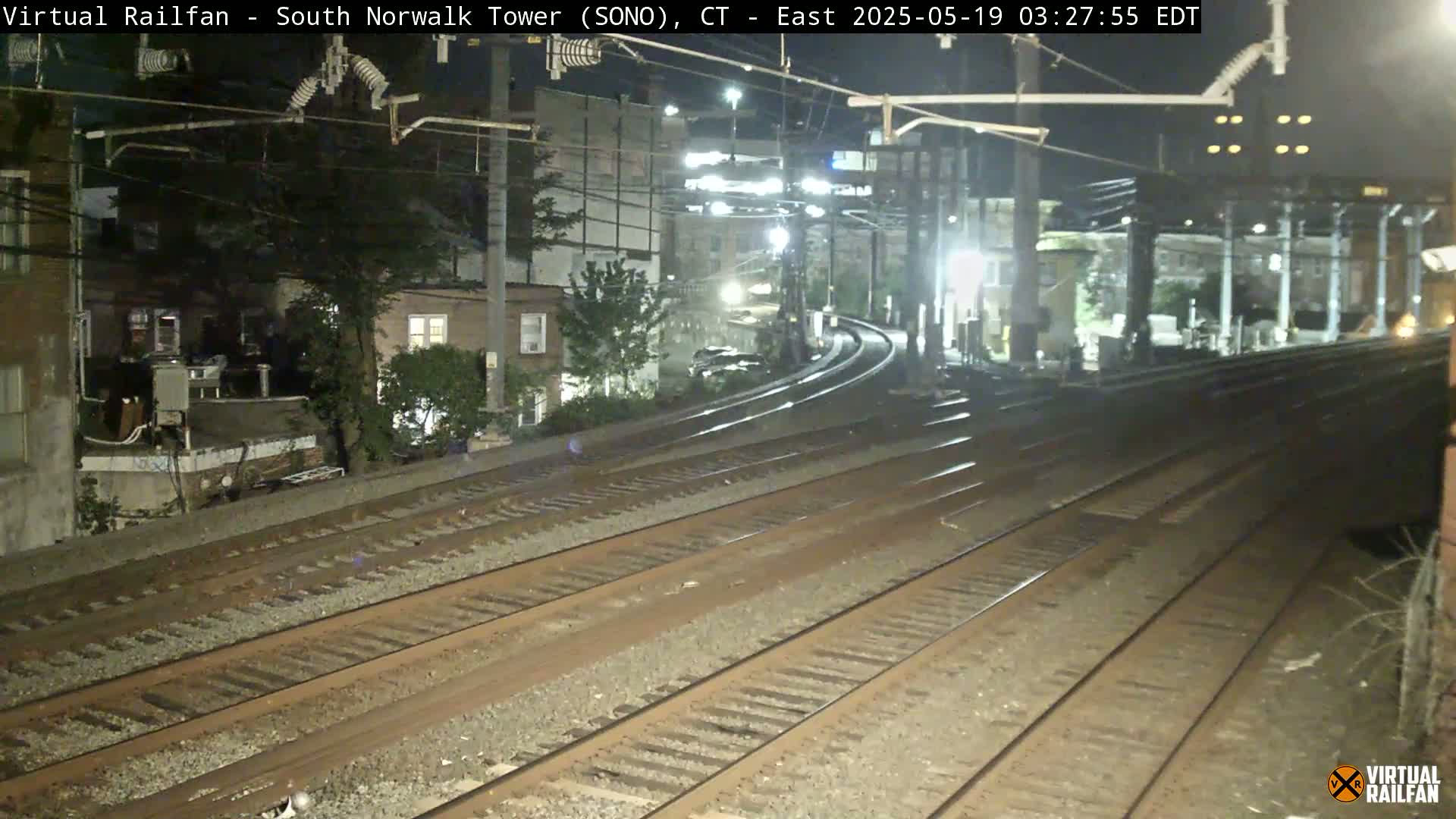 A nighttime view of multiple train tracks curving away from the camera, passing beside buildings and trees under artificial light.