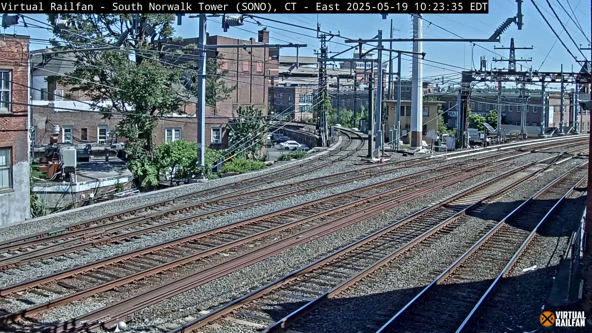 Multiple train tracks curve through a sunny urban area with brick buildings and trees.