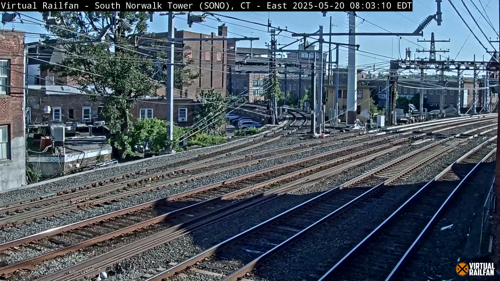 Multiple train tracks extend into the distance, past buildings and trees under a sunny sky.