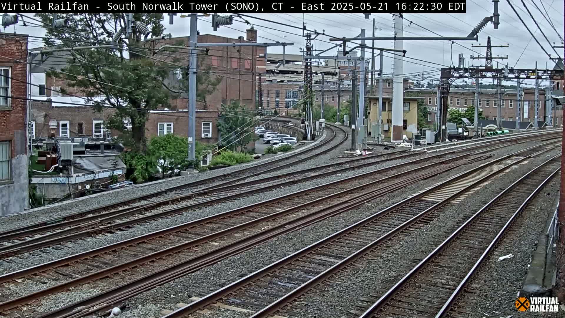 Multiple train tracks curve gently to the right, passing in front of brick buildings under an overcast sky.