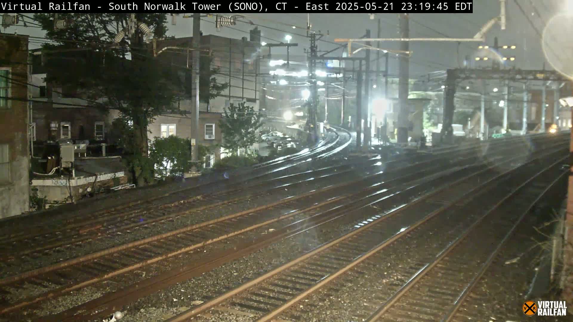 Wet railroad tracks curve through a nighttime cityscape under rainy conditions.