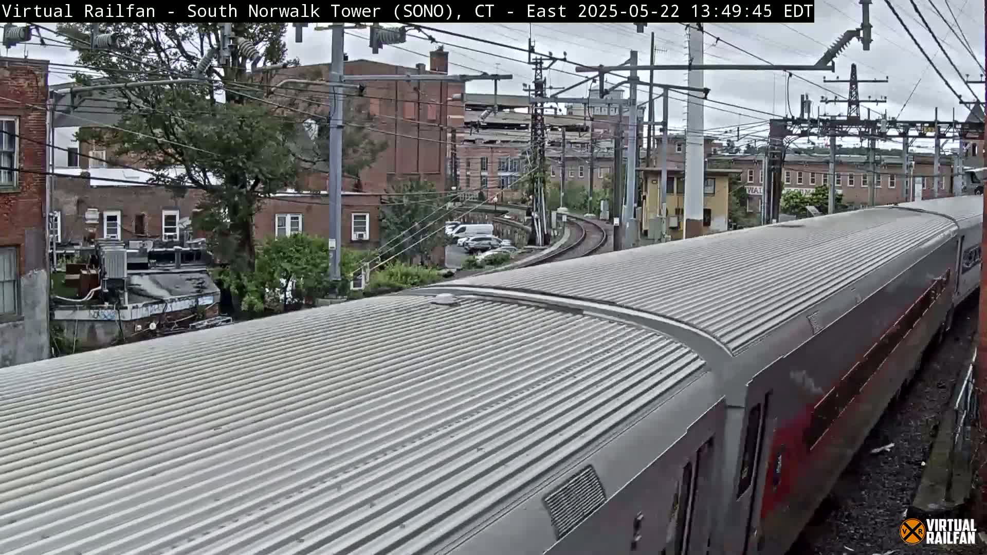A long train with a ribbed metal roof is passing through a town under an overcast sky.