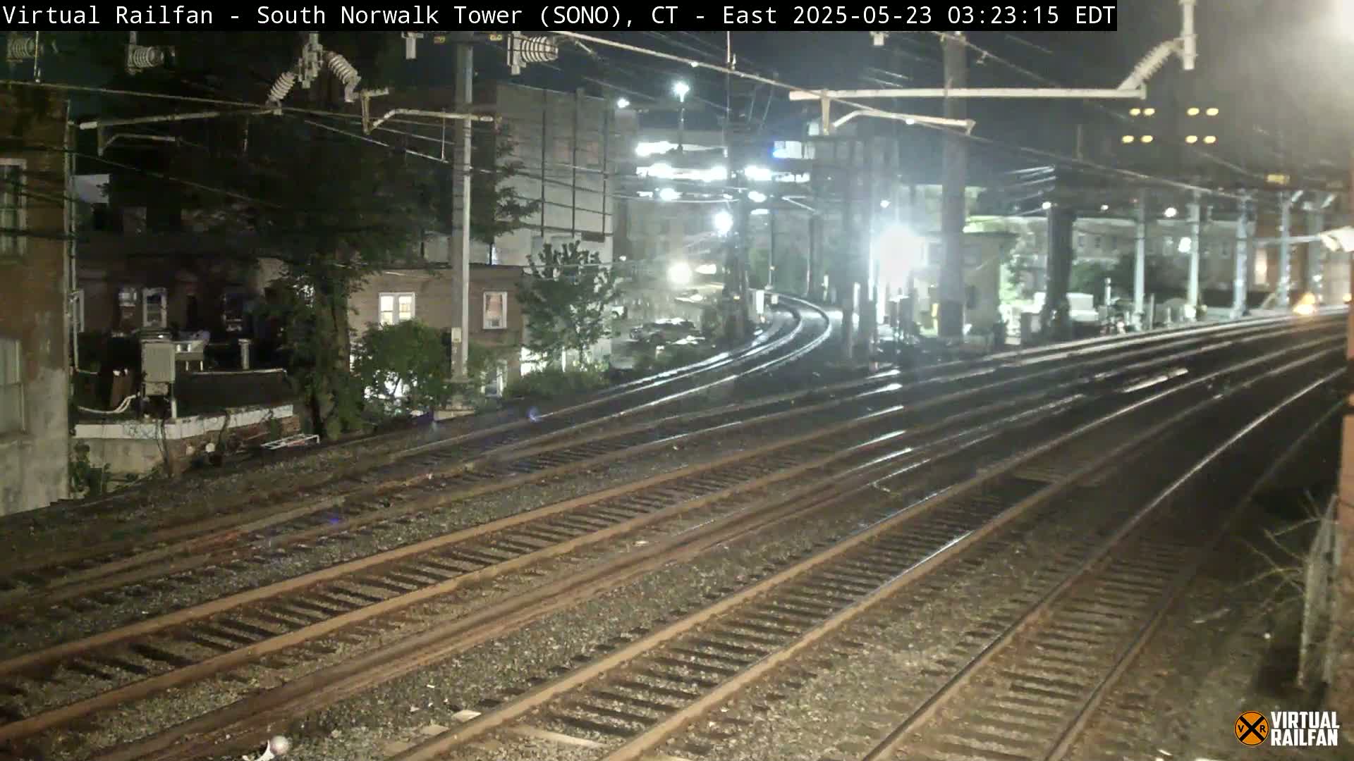 At night, several train tracks curve away from the viewer toward buildings under dim lighting.