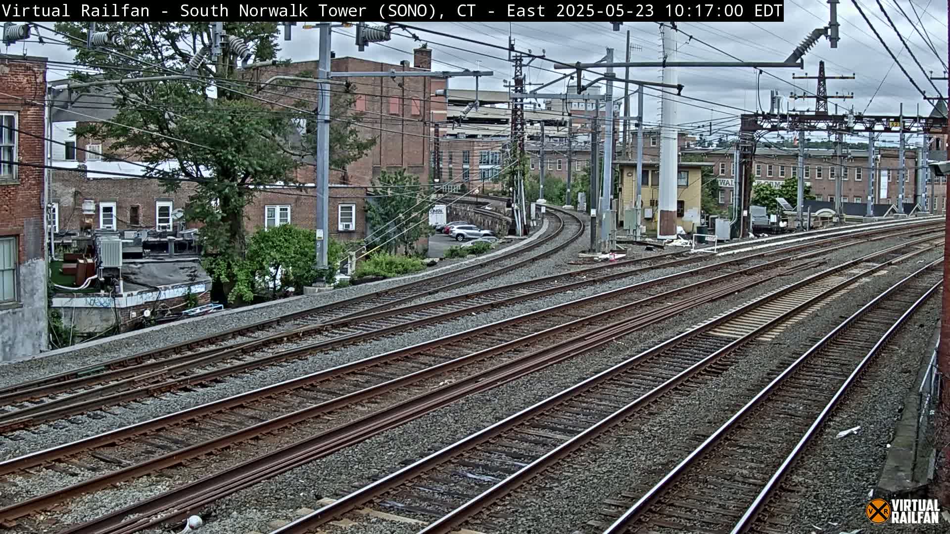 Several sets of train tracks curve gently away from the camera, passing brick buildings and trees under an overcast sky.
