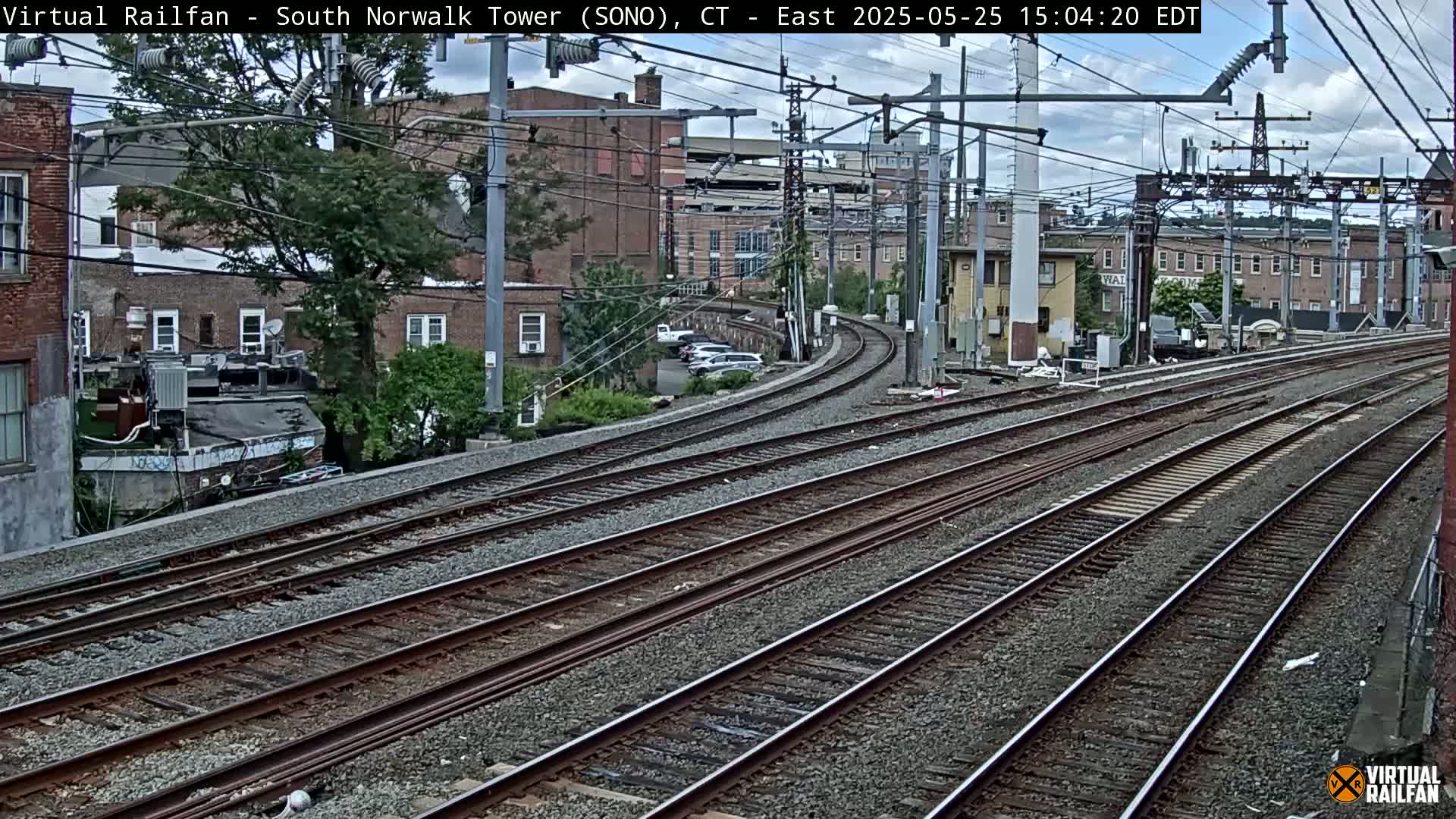 Several sets of train tracks curve gently to the right, passing in front of brick buildings under a partly cloudy sky.