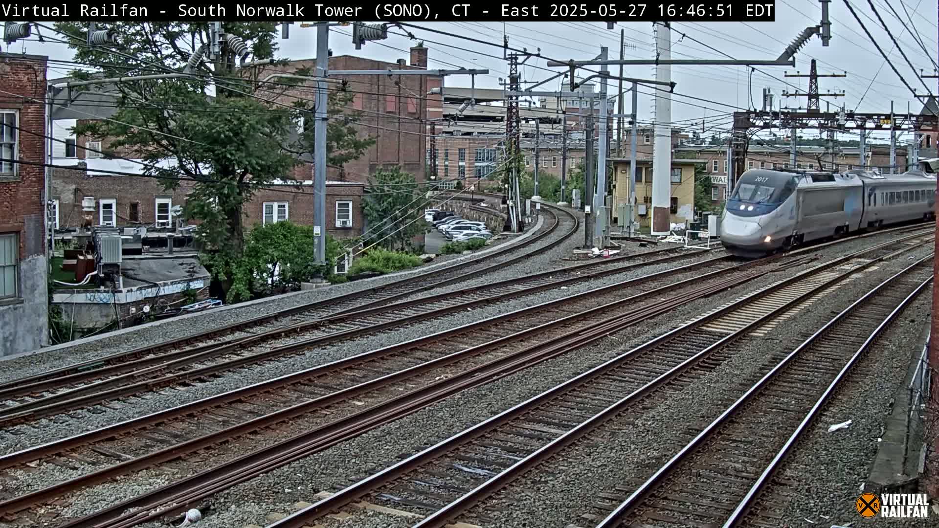 A high-speed train passes by on a curved section of railroad tracks on an overcast day, with several buildings and parked cars visible in the background.