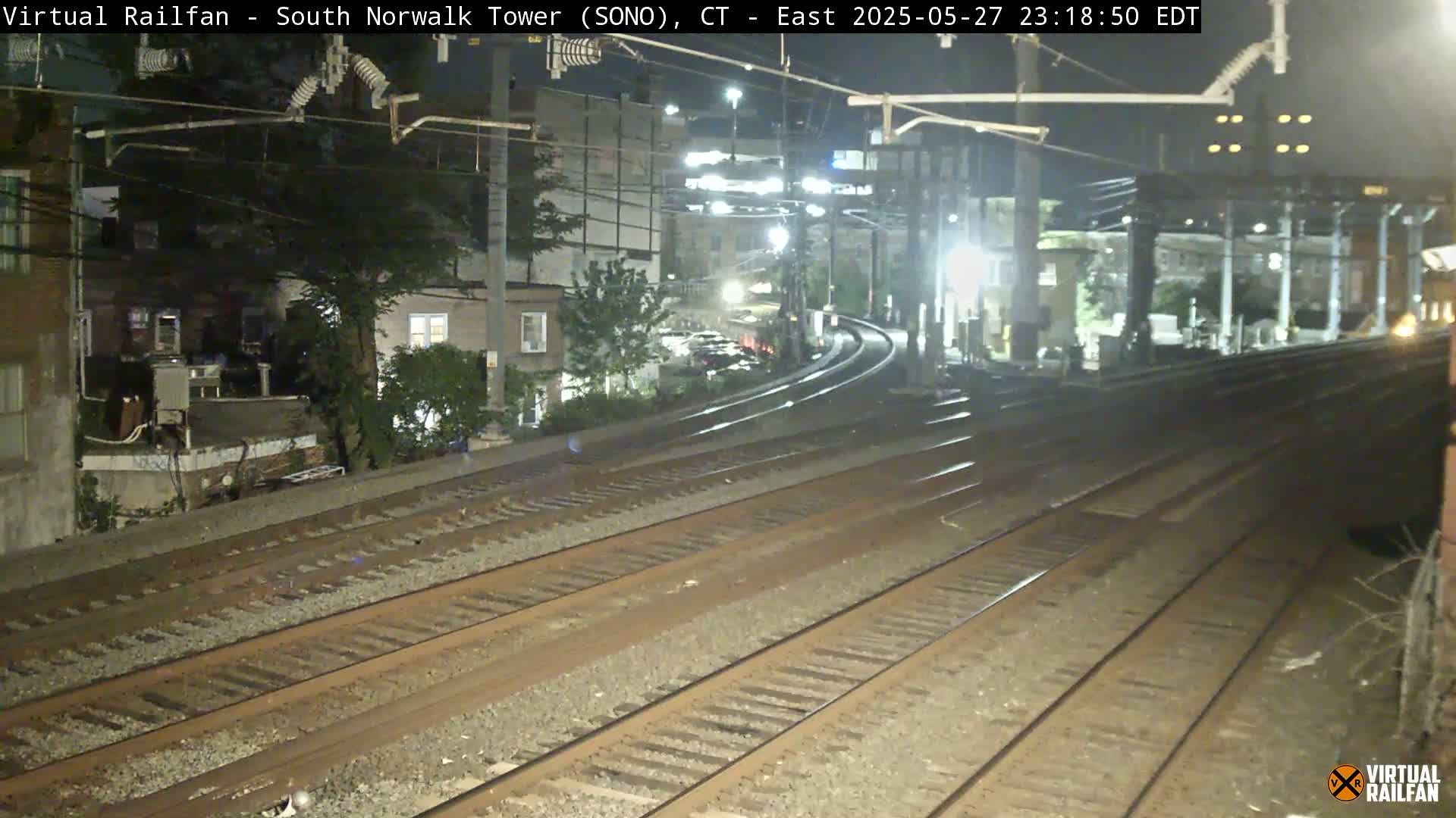 At night, several sets of train tracks curve gently toward a lighted train platform, past buildings and trees.