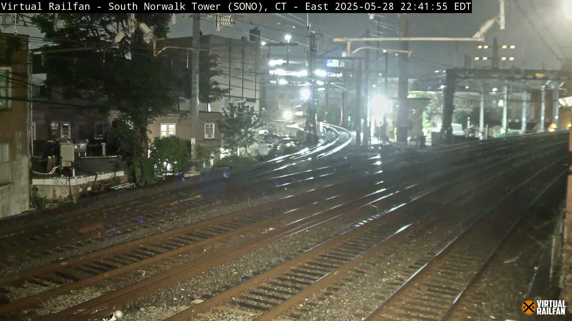 A nighttime view of rain-slicked train tracks curving away from the camera, past residential buildings under partly cloudy skies.