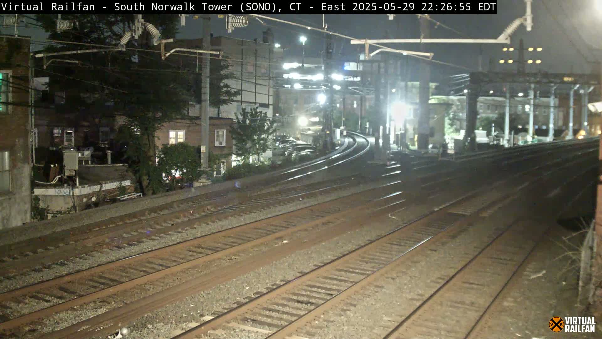 A nighttime view of multiple train tracks curving towards a station, with buildings and trees visible in the background under dark, clear skies.