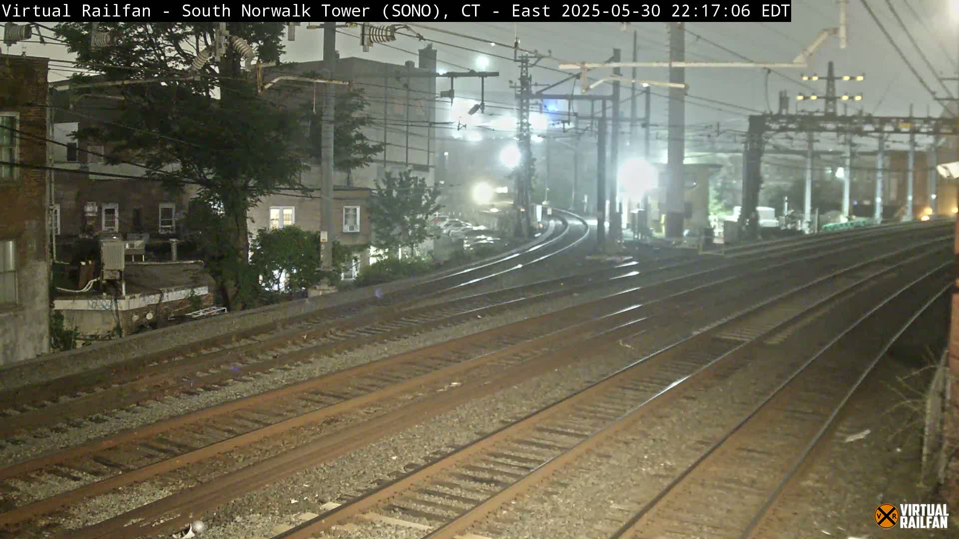 Multiple train tracks diverge in the nighttime mist, passing near buildings and trees under dim lighting.