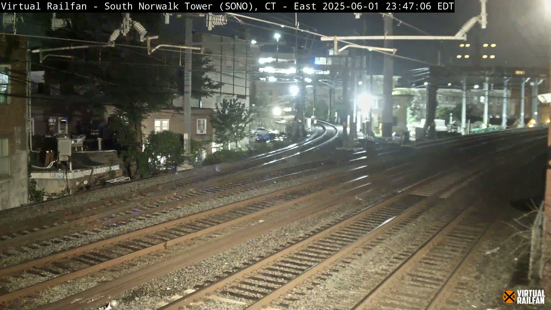 At night, several sets of train tracks curve through a residential area under overhead power lines.