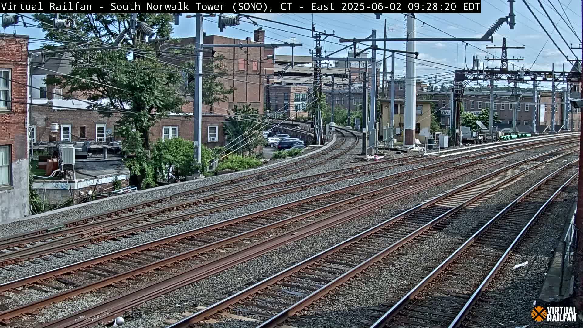 Multiple sets of train tracks curve away from the camera, passing in front of buildings and a parking lot under a partly cloudy sky.
