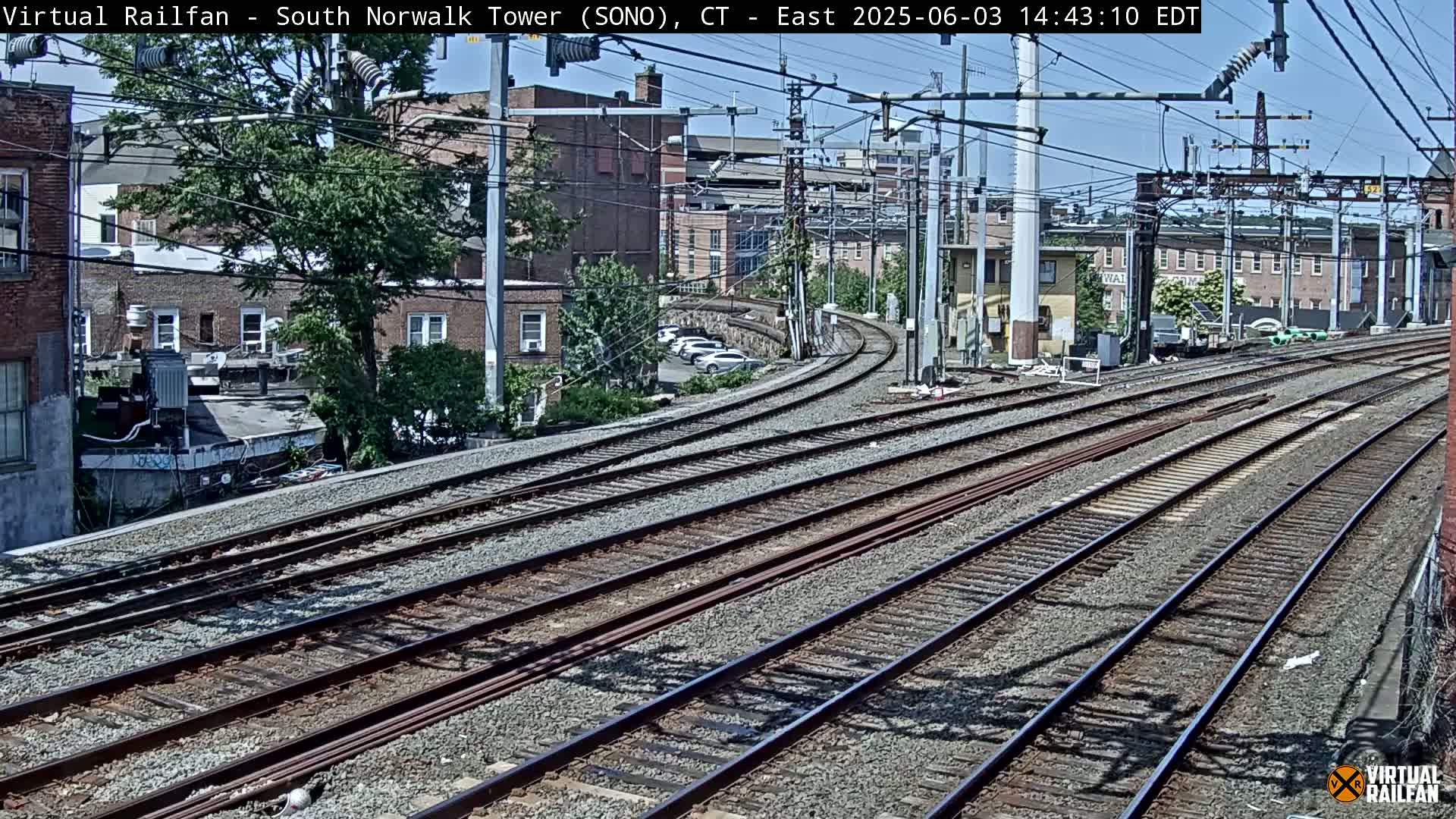 Several sets of train tracks curve gently away from the camera, past brick buildings and leafy trees under a sunny sky.