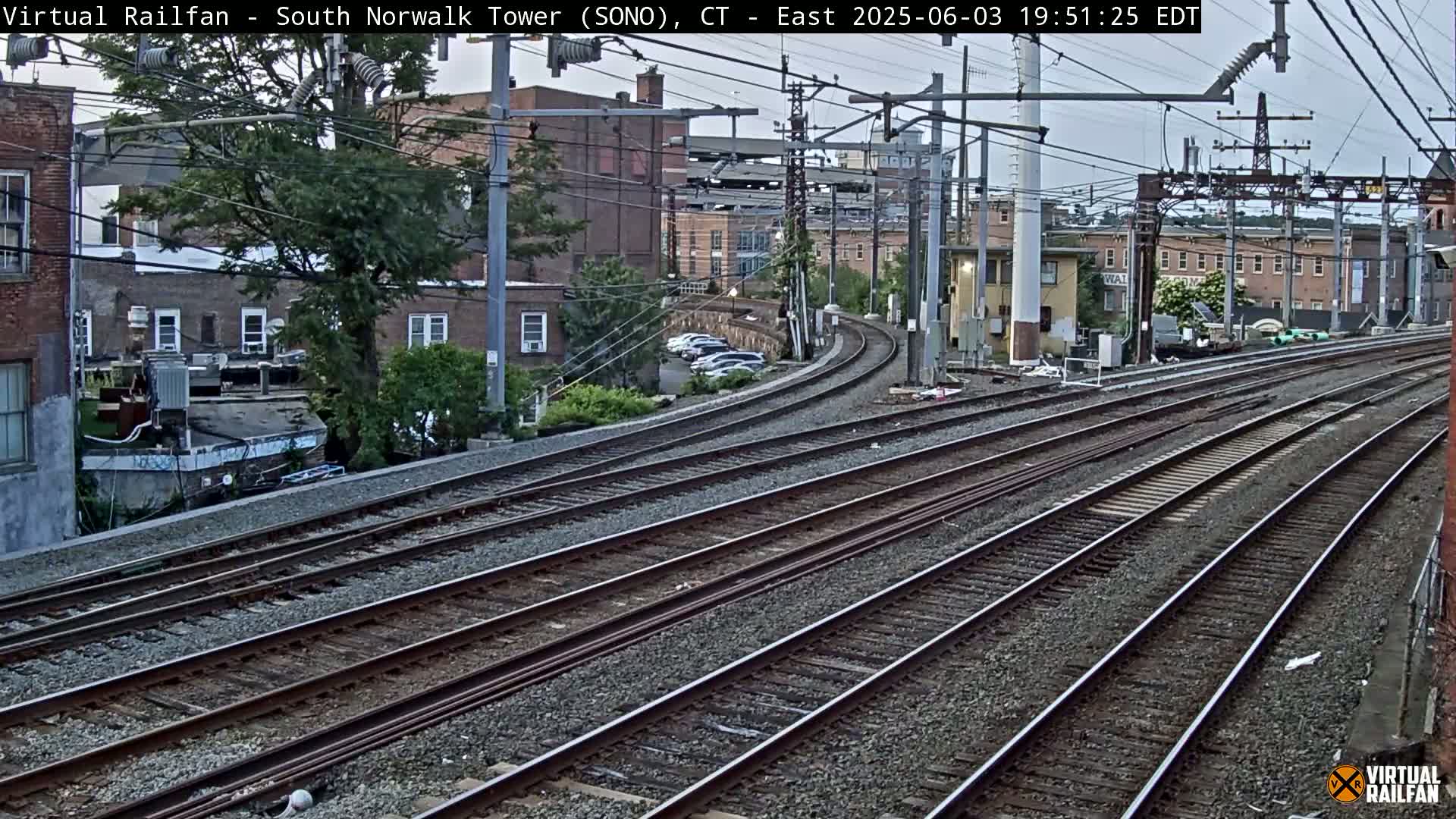 Several sets of train tracks curve away from the viewer, passing in front of buildings and a few parked cars under an overcast sky.