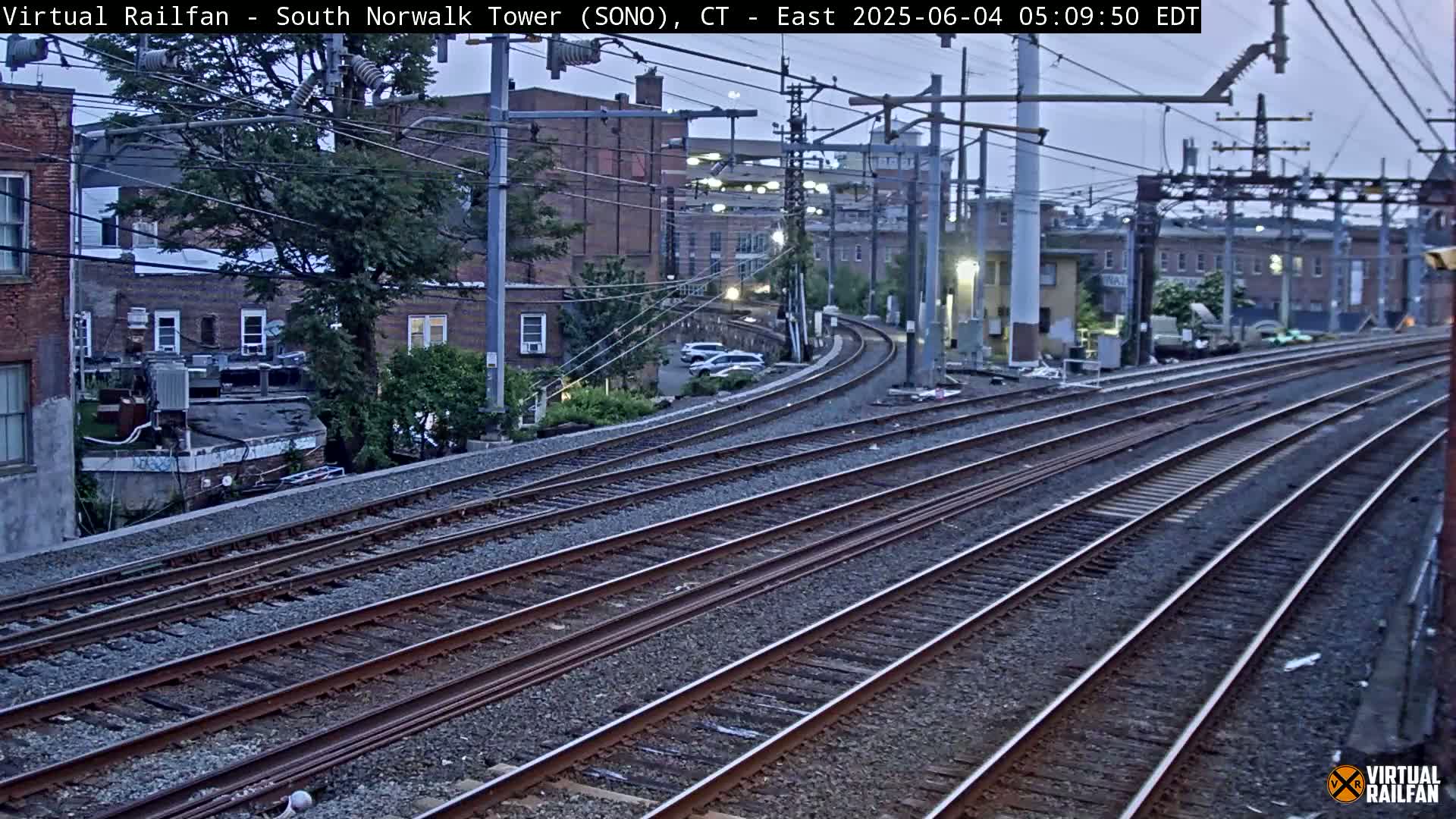 Multiple sets of train tracks curve gently away from the camera, passing in front of buildings and trees under an overcast sky.