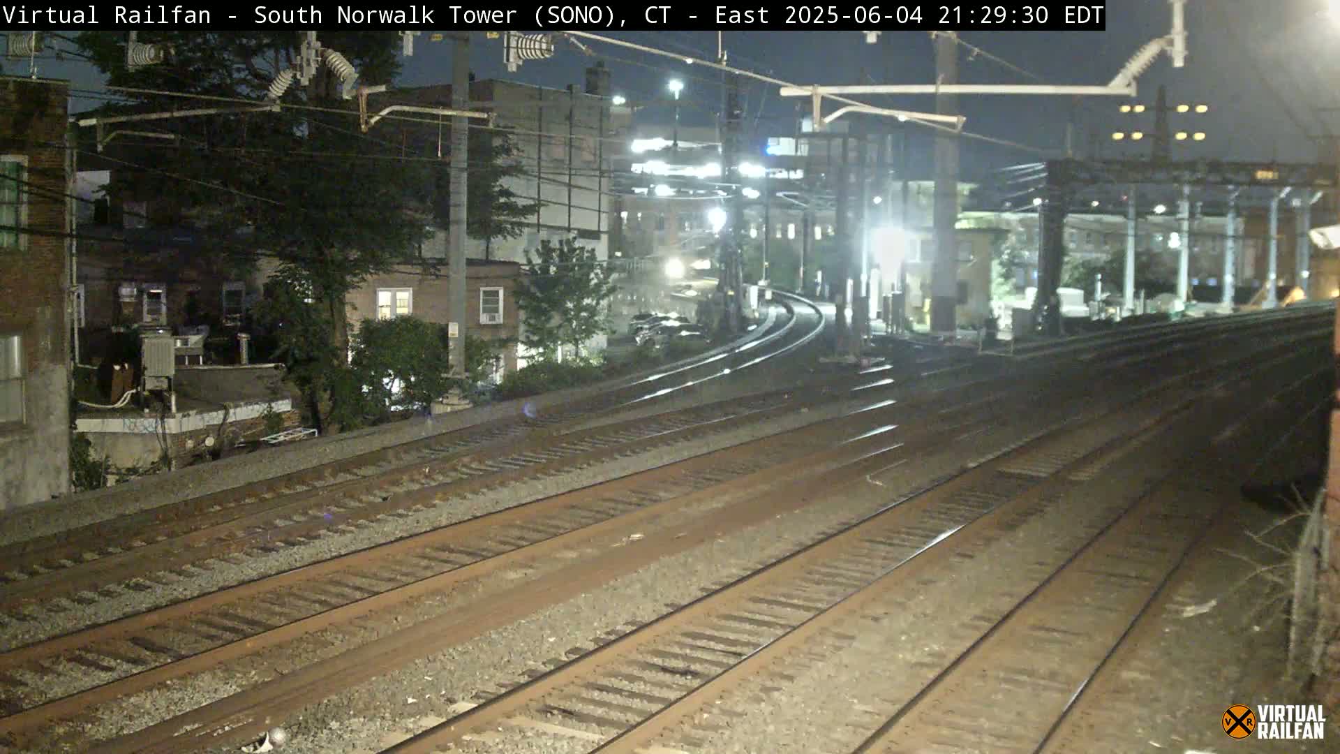 At night, several sets of train tracks extend into the distance, past buildings and trees under a dark sky.