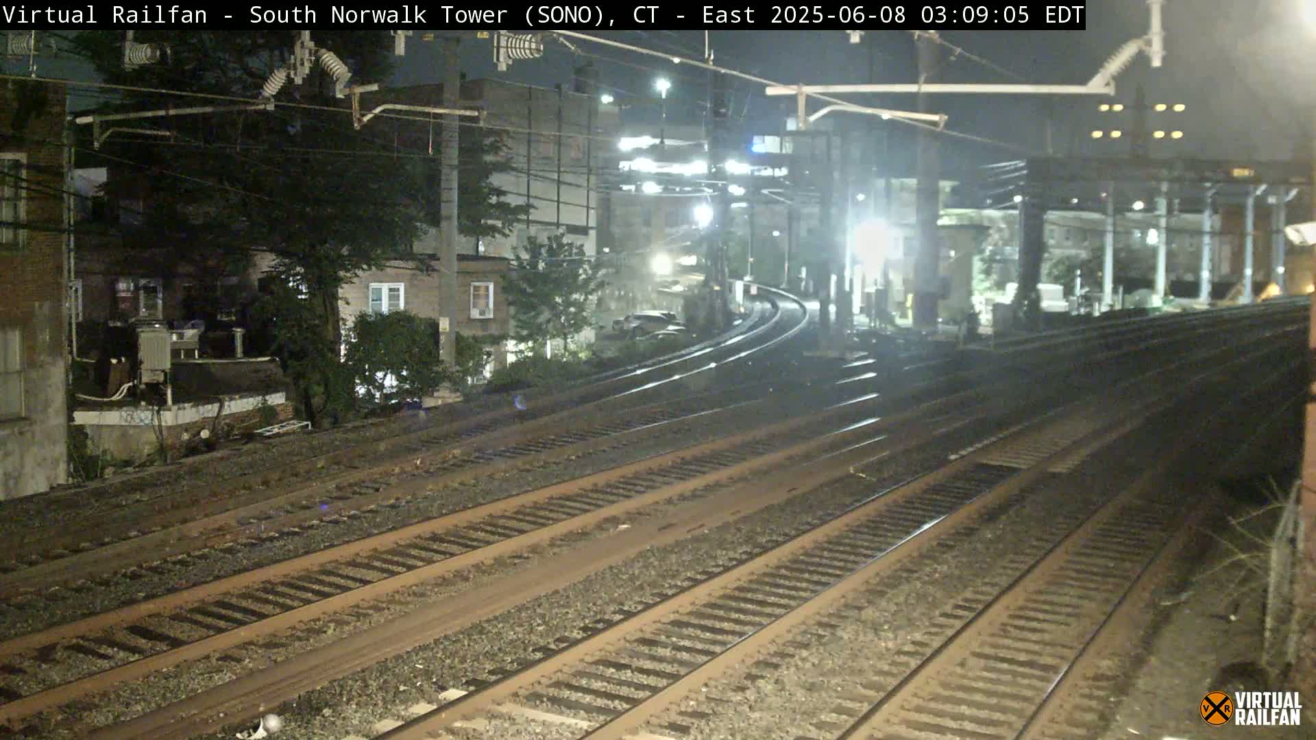 At night, several train tracks curve away from the viewer, passing alongside residential buildings under overhead power lines.