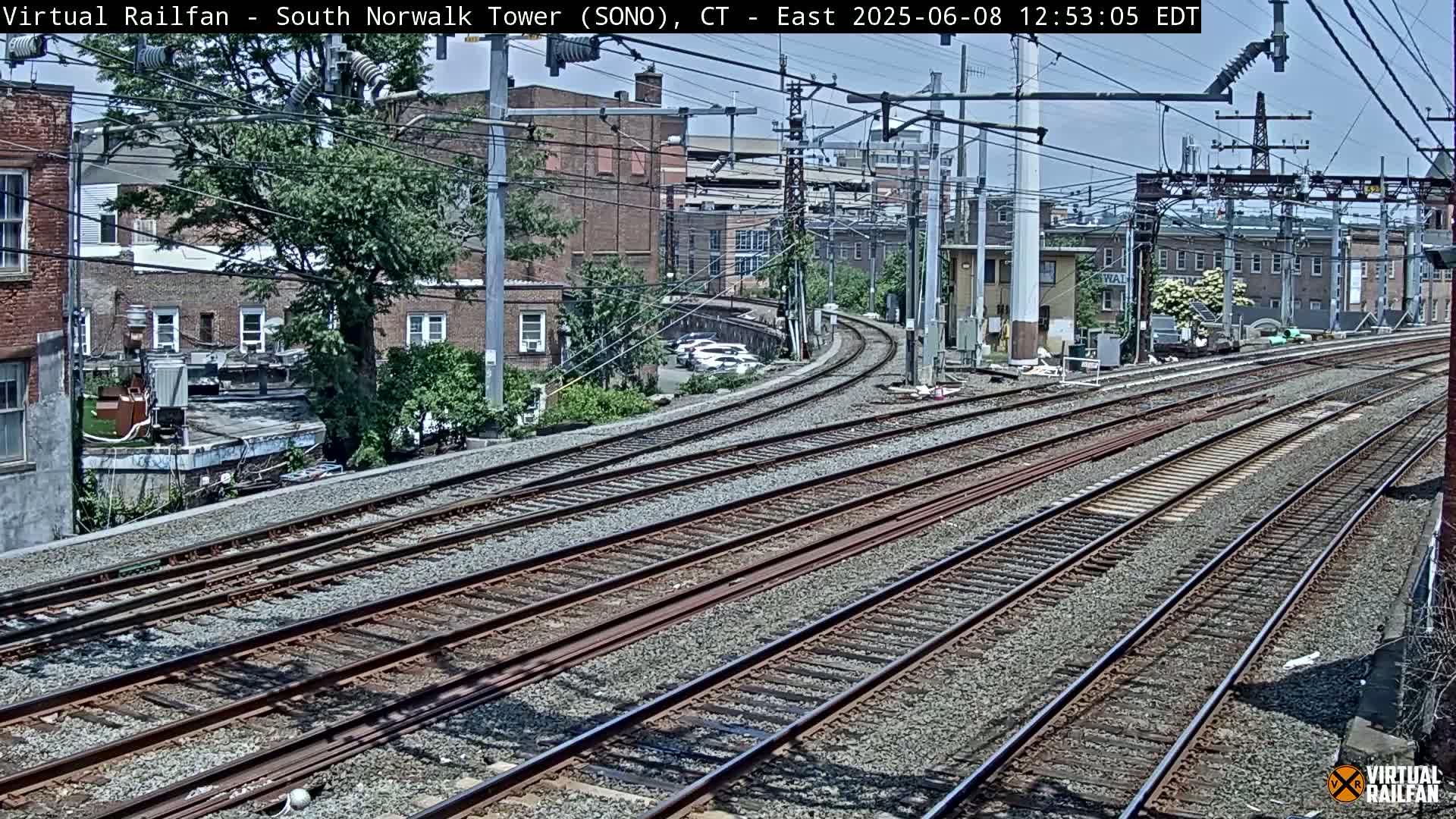 Multiple sets of train tracks curve away from the viewer, passing in front of buildings and trees under a partly sunny sky.