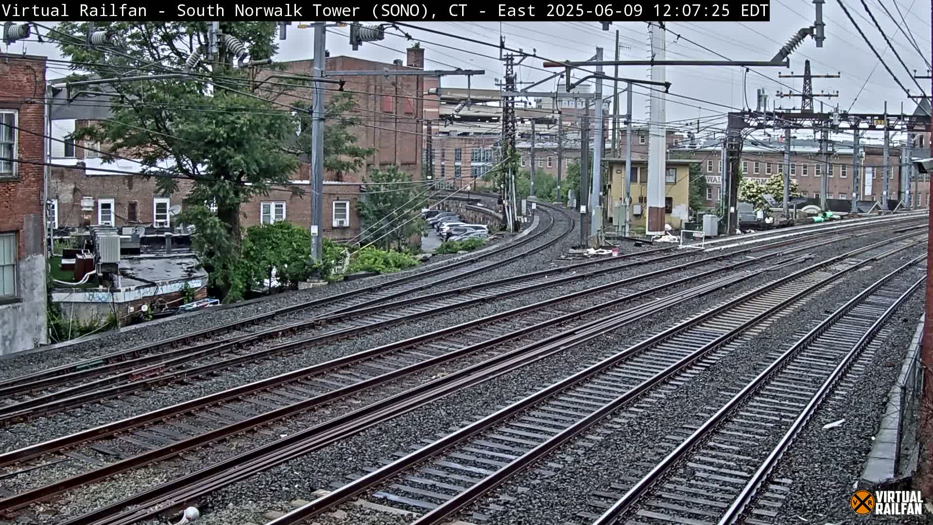 Multiple sets of train tracks curve away from the camera, passing brick buildings and lush greenery under an overcast sky.