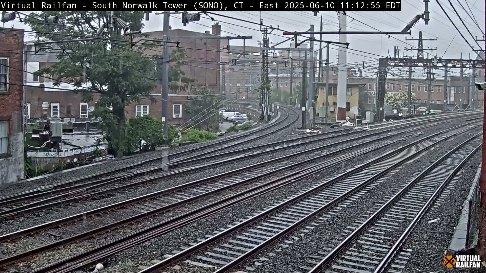 Several sets of train tracks curve away from the camera under an overcast sky, passing by buildings in a rainy urban environment.