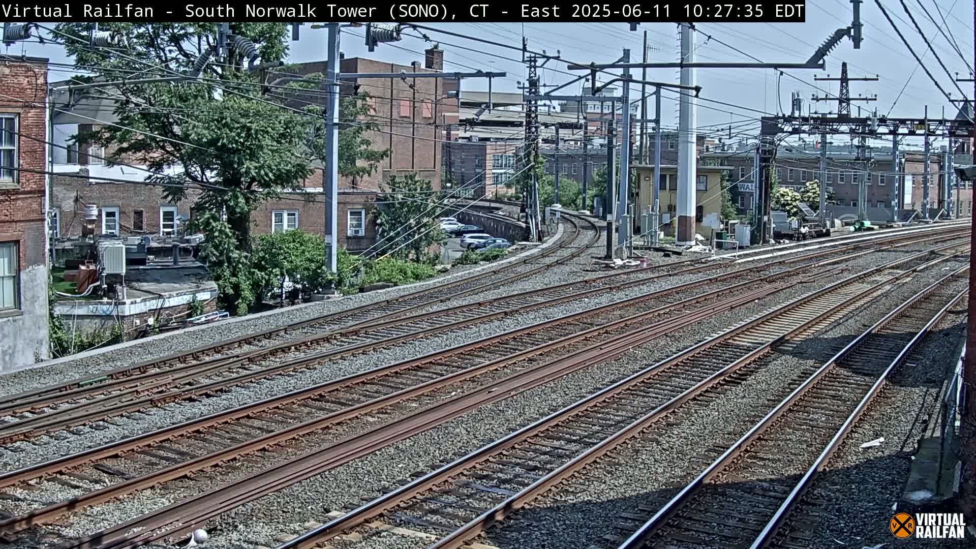 Several sets of train tracks curve gently through a sunny urban area, past brick buildings and trees.
