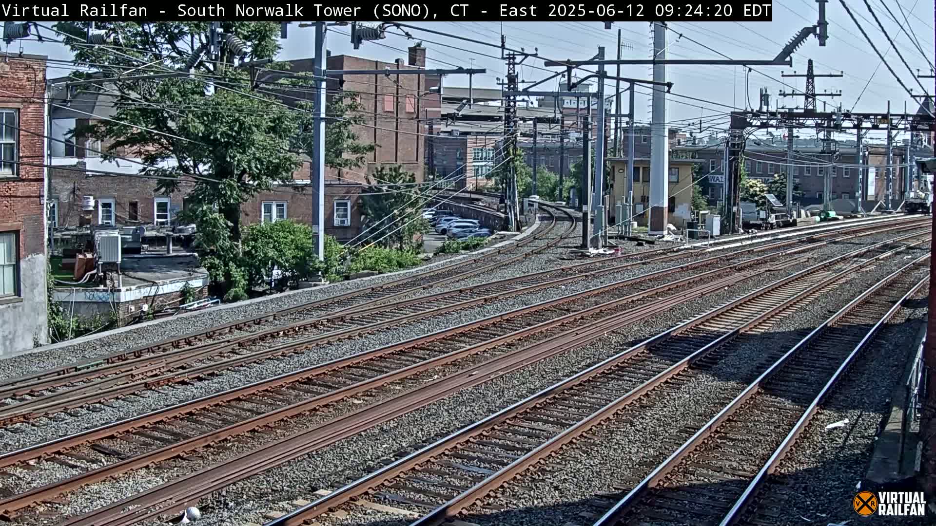 A sunny day view of multiple sets of train tracks curving away from the camera, past buildings and some parked cars.
