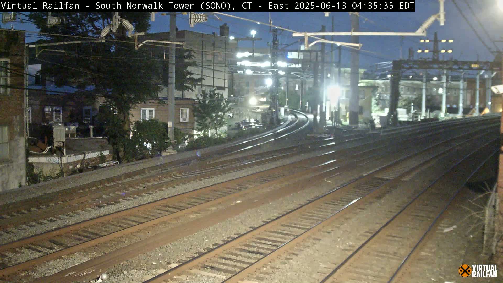 Multiple train tracks diverge at night under a mostly clear sky, passing near residential buildings.