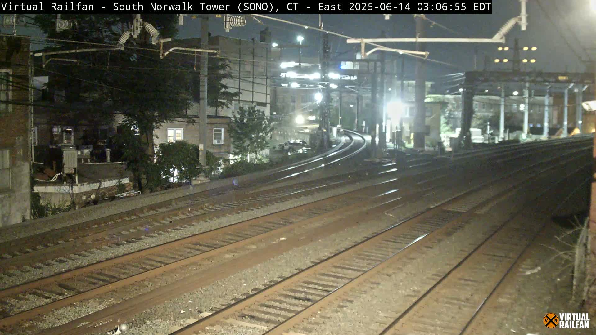 At night, several sets of train tracks curve gently toward a platform next to buildings under a dark sky.