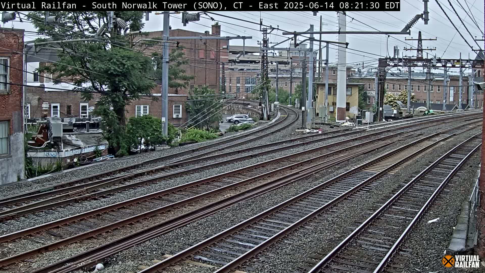 Multiple sets of train tracks curve gently to the left, passing brick buildings and lush green trees under an overcast sky.
