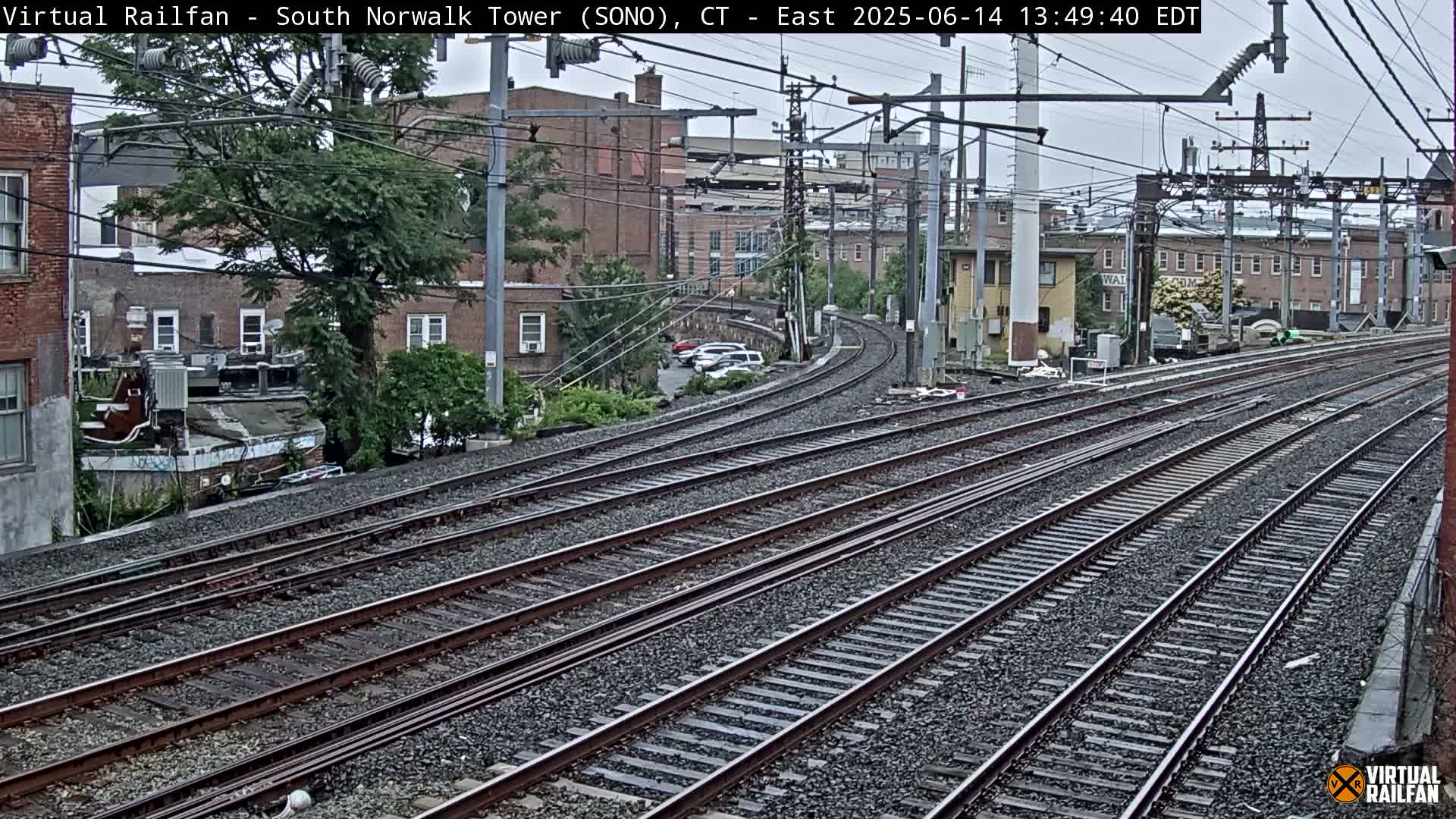 Multiple sets of train tracks curve gently away from the camera, passing brick buildings and trees under an overcast sky.