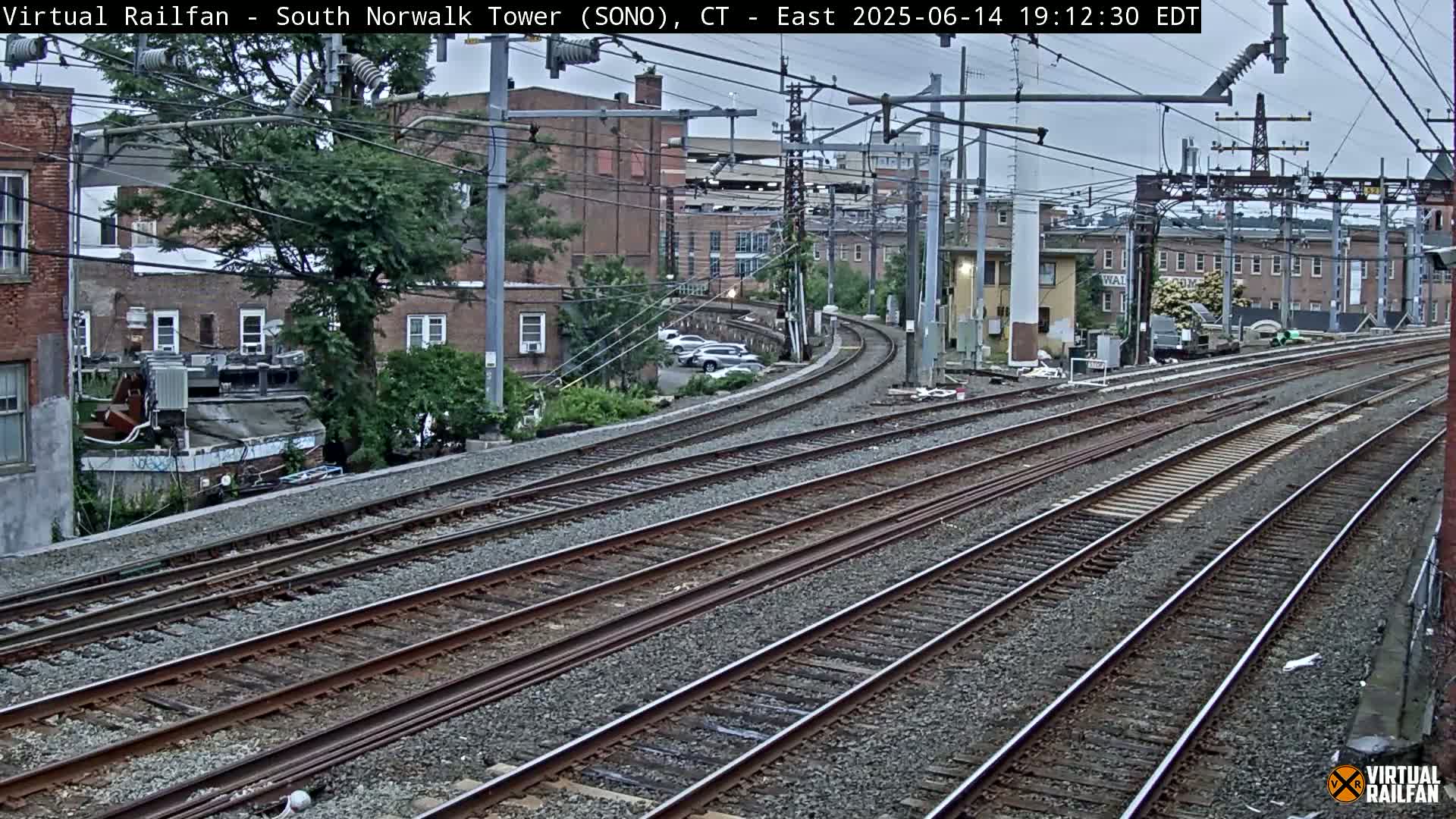 Multiple sets of train tracks curve gently away from the camera, passing brick buildings and trees under an overcast sky.