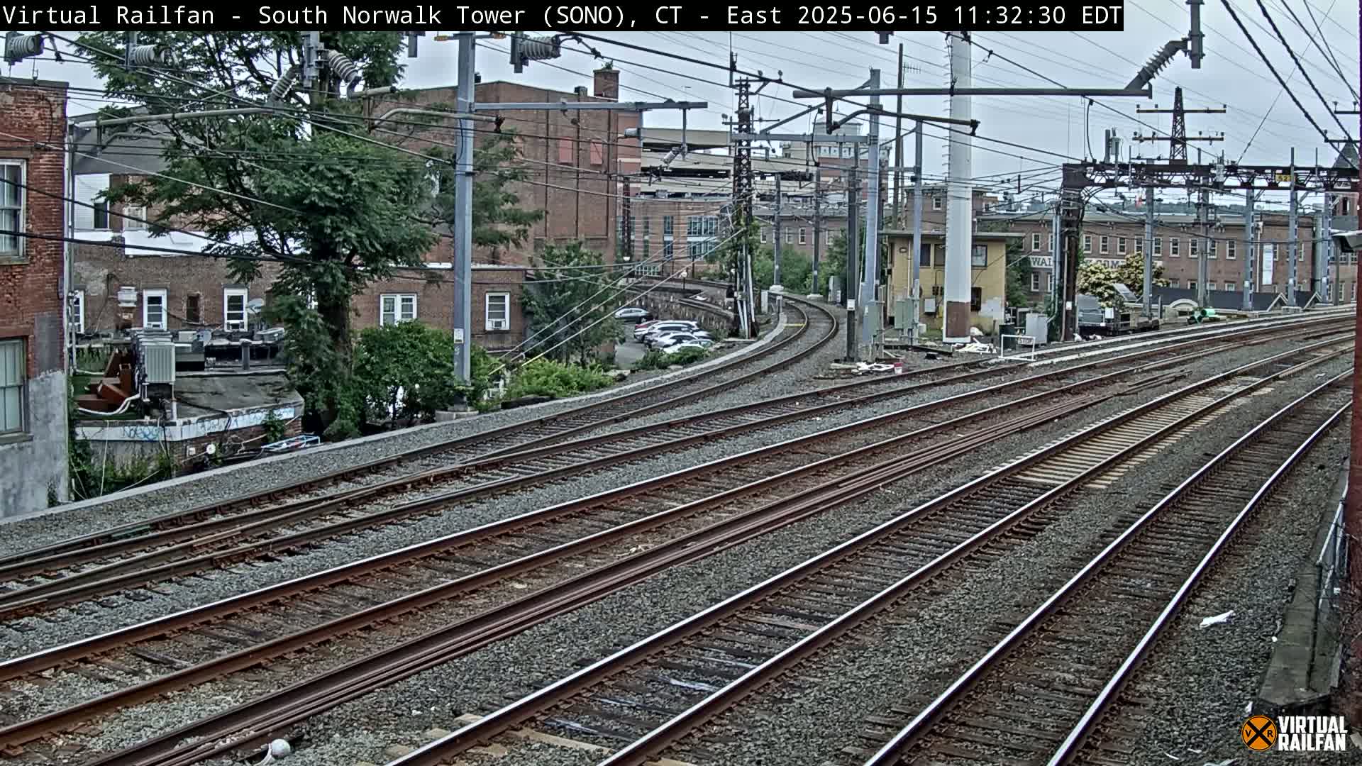 Several sets of train tracks curve gently to the left, passing brick buildings and a tree under an overcast sky.