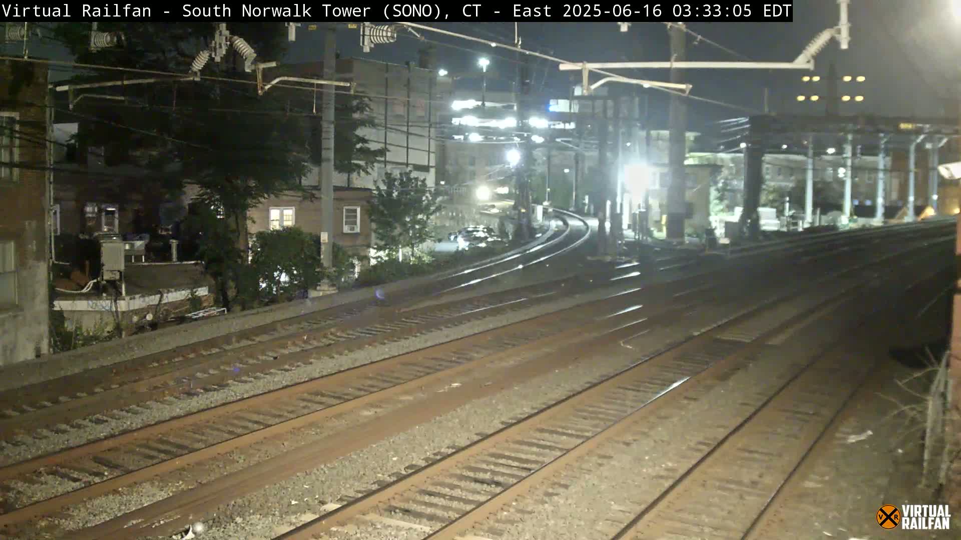A nighttime view of multiple train tracks curving away from the camera, passing by buildings and trees under artificial light.