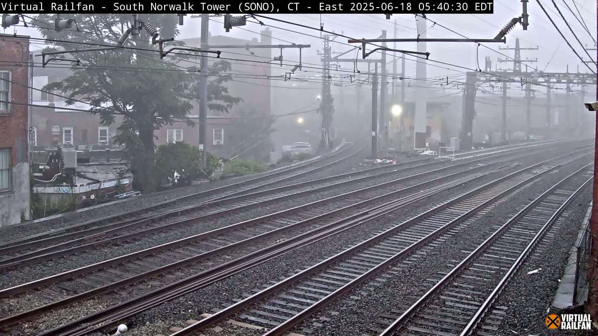 Multiple sets of train tracks recede into a misty, rainy scene, past buildings and overhead power lines.