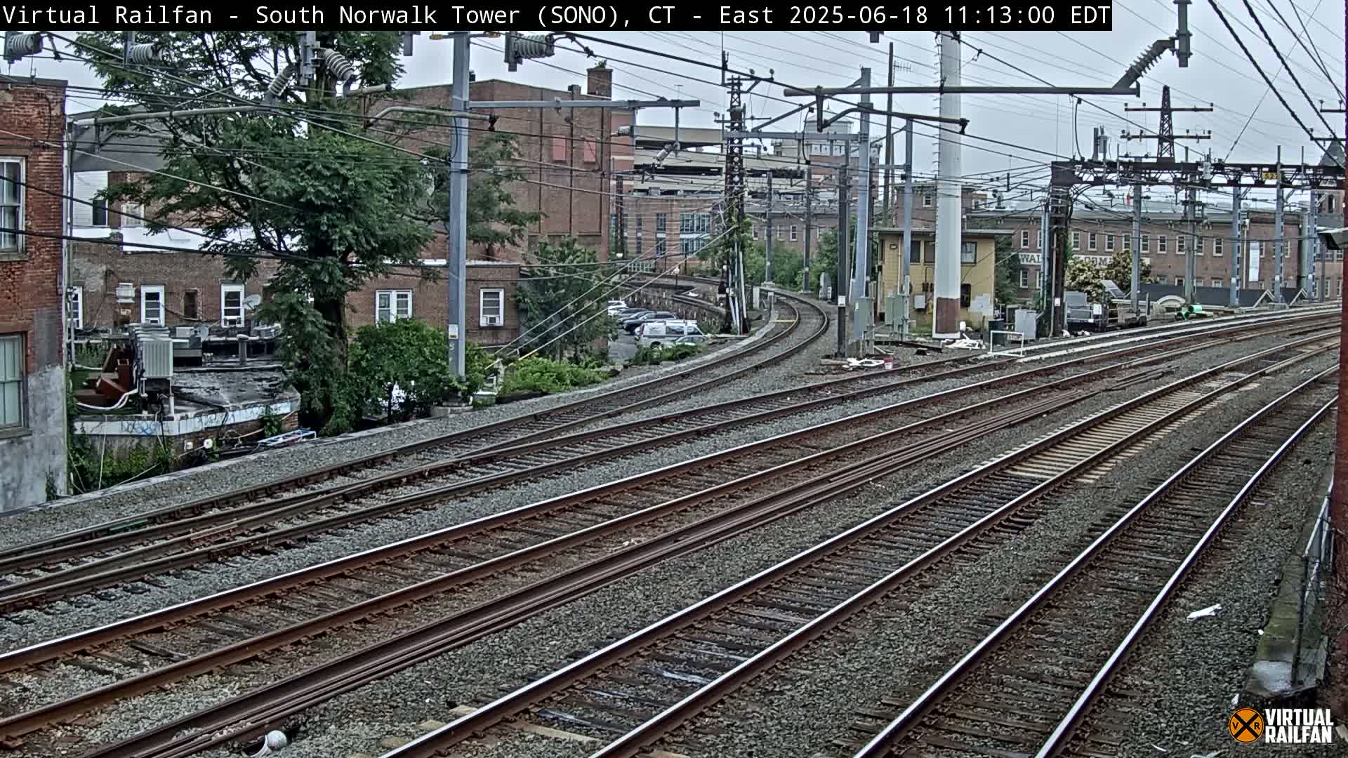 Several parallel train tracks curve gently to the left, passing alongside brick buildings and trees under an overcast sky.