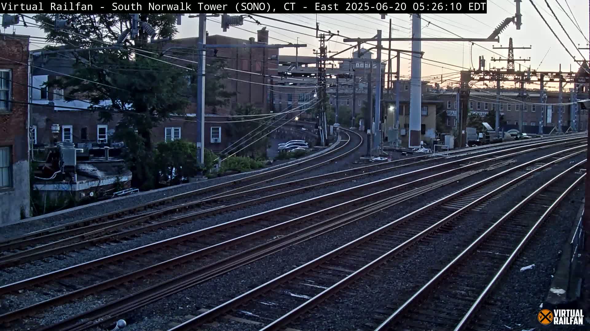 Multiple train tracks curve gently through an urban area at twilight, passing by buildings and a large tree.