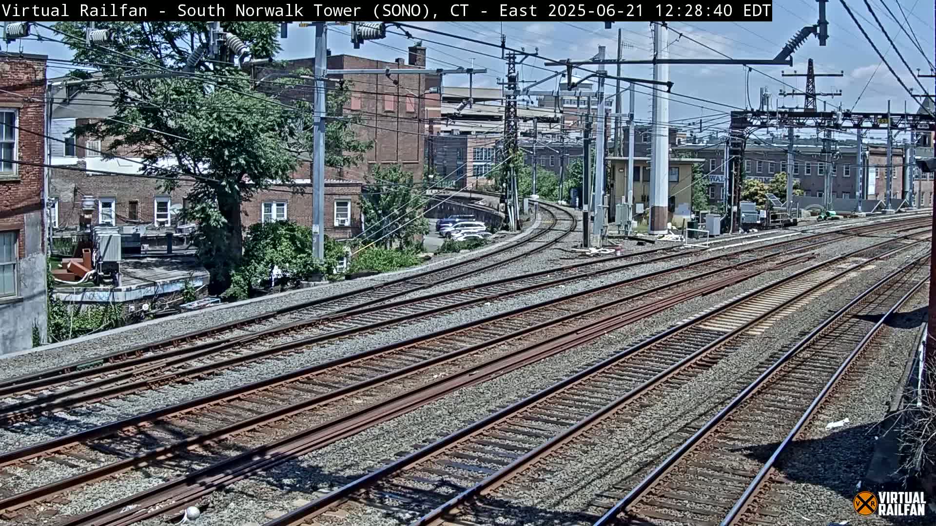A sunny day view of multiple train tracks curving through a city, with buildings and trees visible in the background.