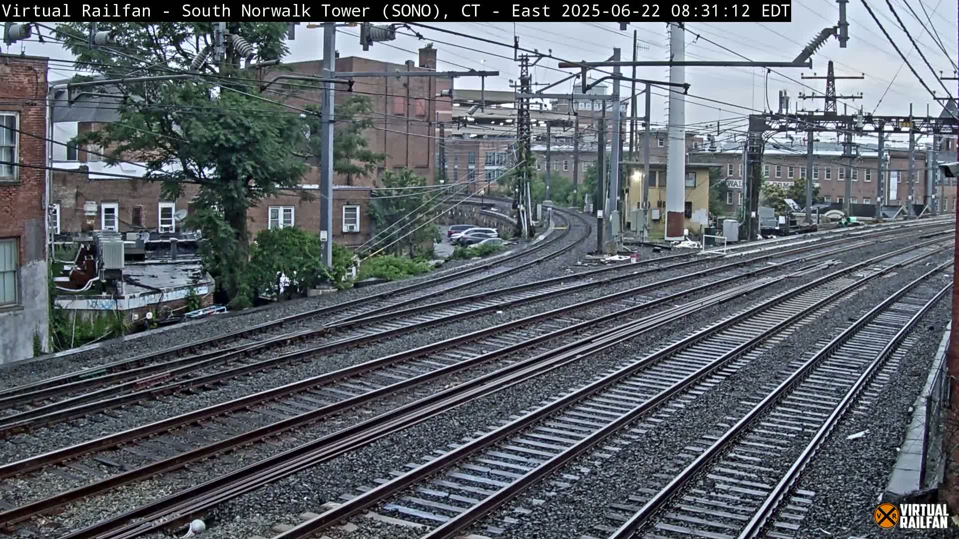 Several sets of train tracks curve gently away from the viewer, past buildings and trees under an overcast sky.