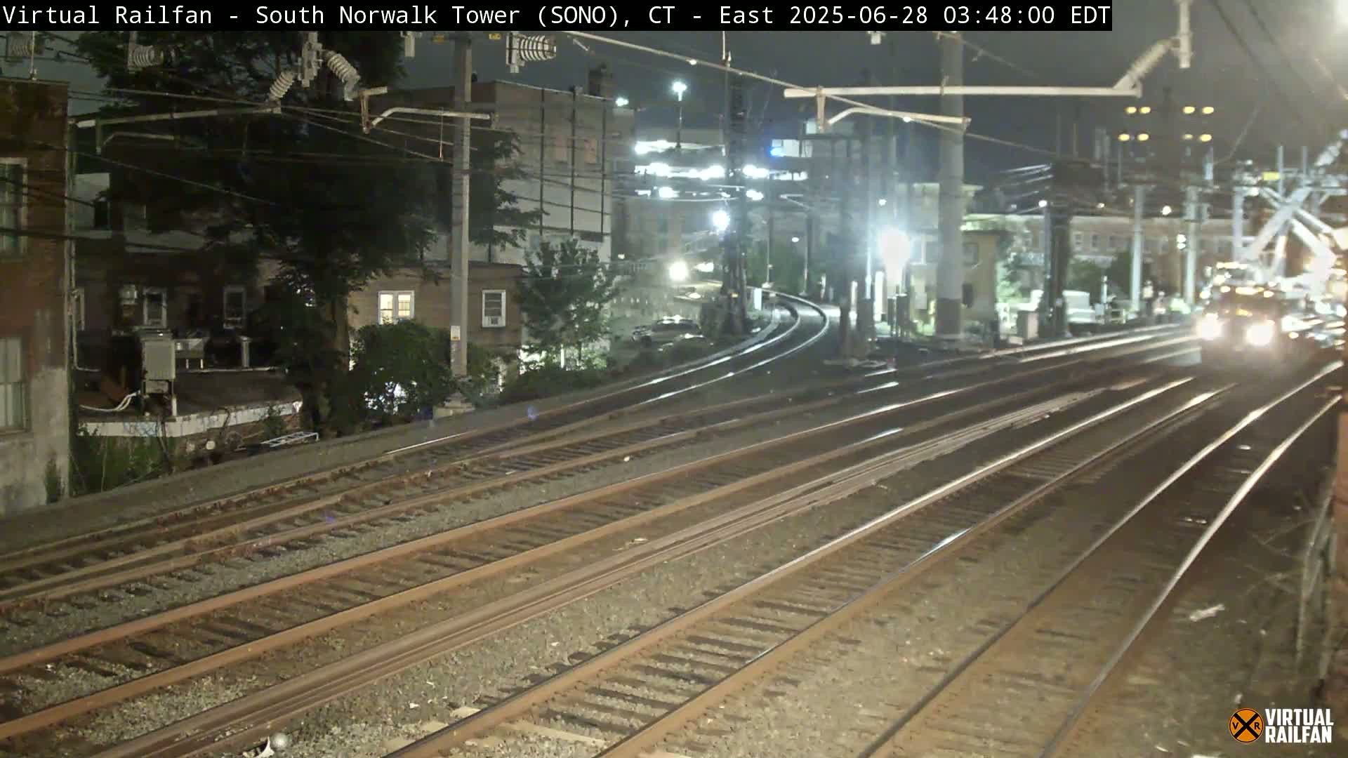 A train approaches at night on multiple train tracks that curve past buildings and trees under dark, nighttime conditions.