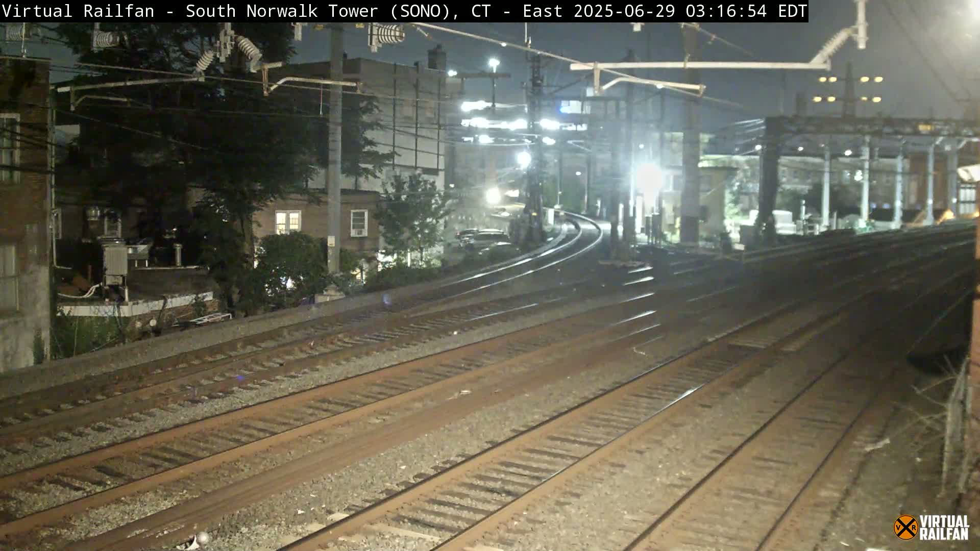 At night, multiple train tracks curve gently to the left, passing near buildings and trees under overhead electrical wires.