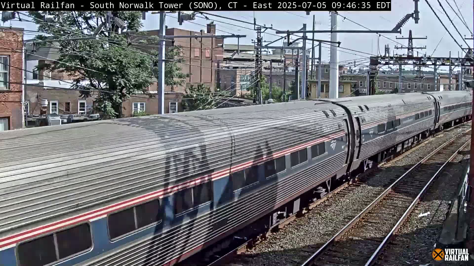A long, silver passenger train passes by on a sunny day, alongside brick buildings and overhead power lines.