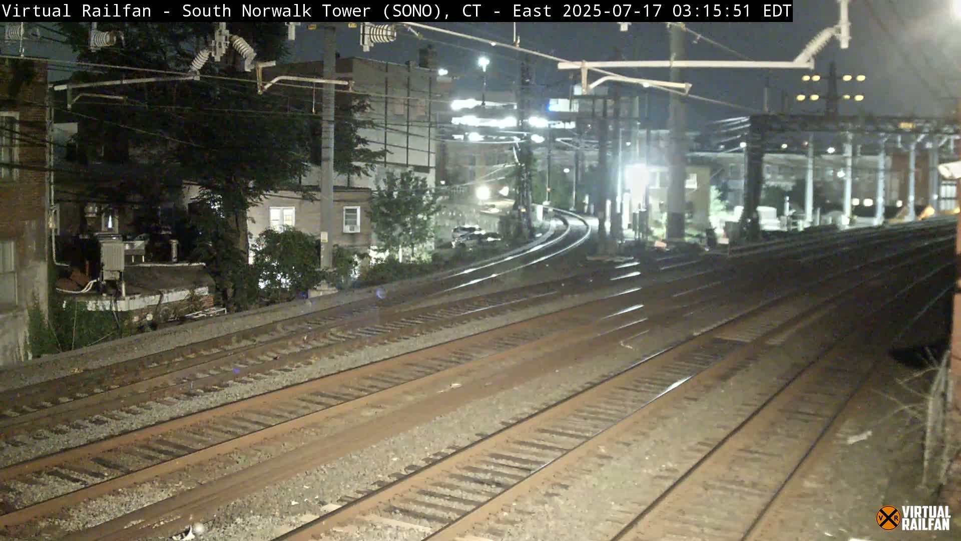 At night, multiple train tracks curve away from the viewer toward buildings and trees under dark skies.