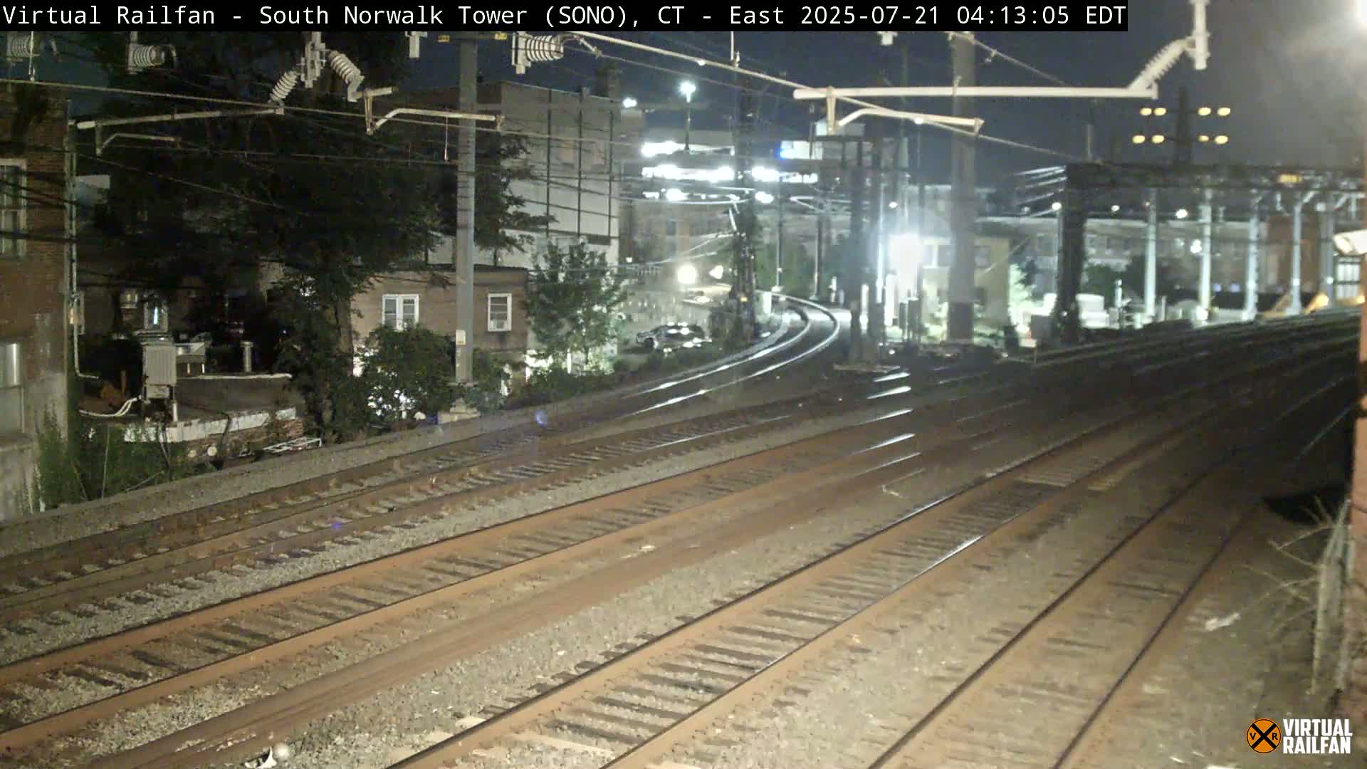 Multiple sets of train tracks curve away from the camera at night, passing near residential buildings and trees under dark skies.